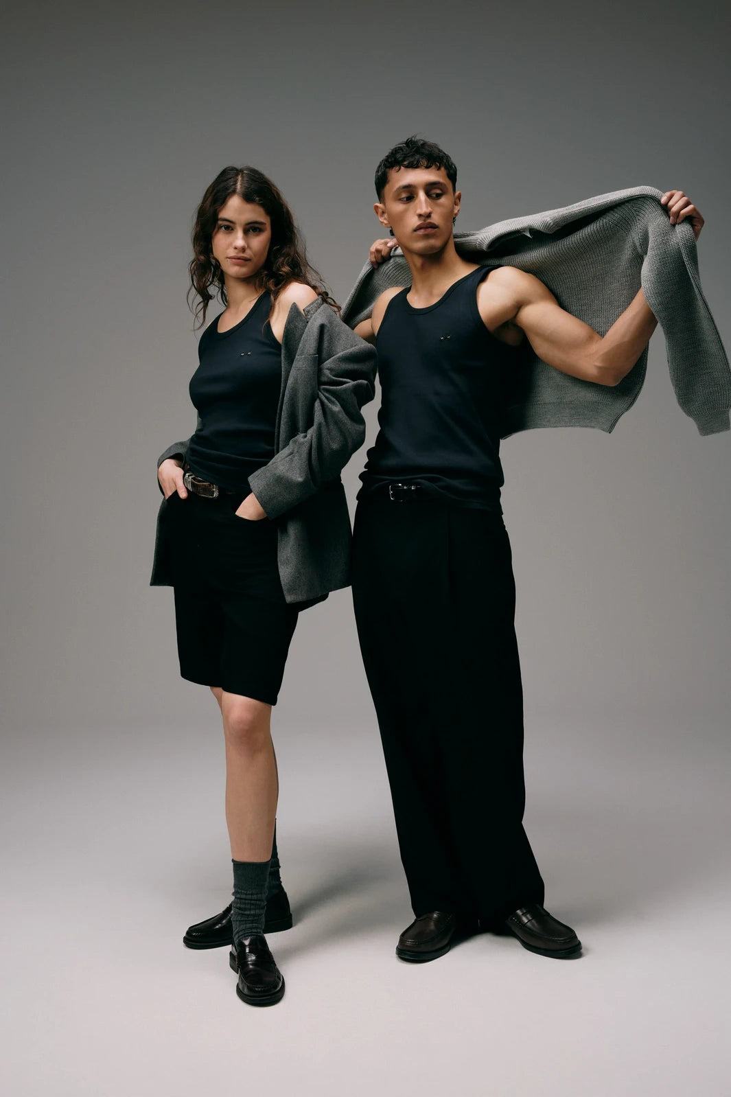 Débardeurs Noirs Tricots Marcel, portés par un homme et une femme avec des vestes grises, vue en pied.