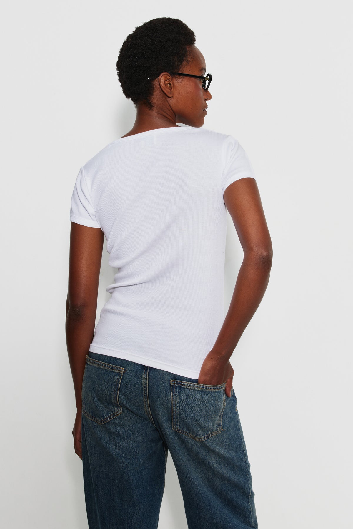 Maillot de corps femme blanc à manches courtes en coton Pima côtelé, porté de dos avec un jean bleu, la tête tournée sur le côté.