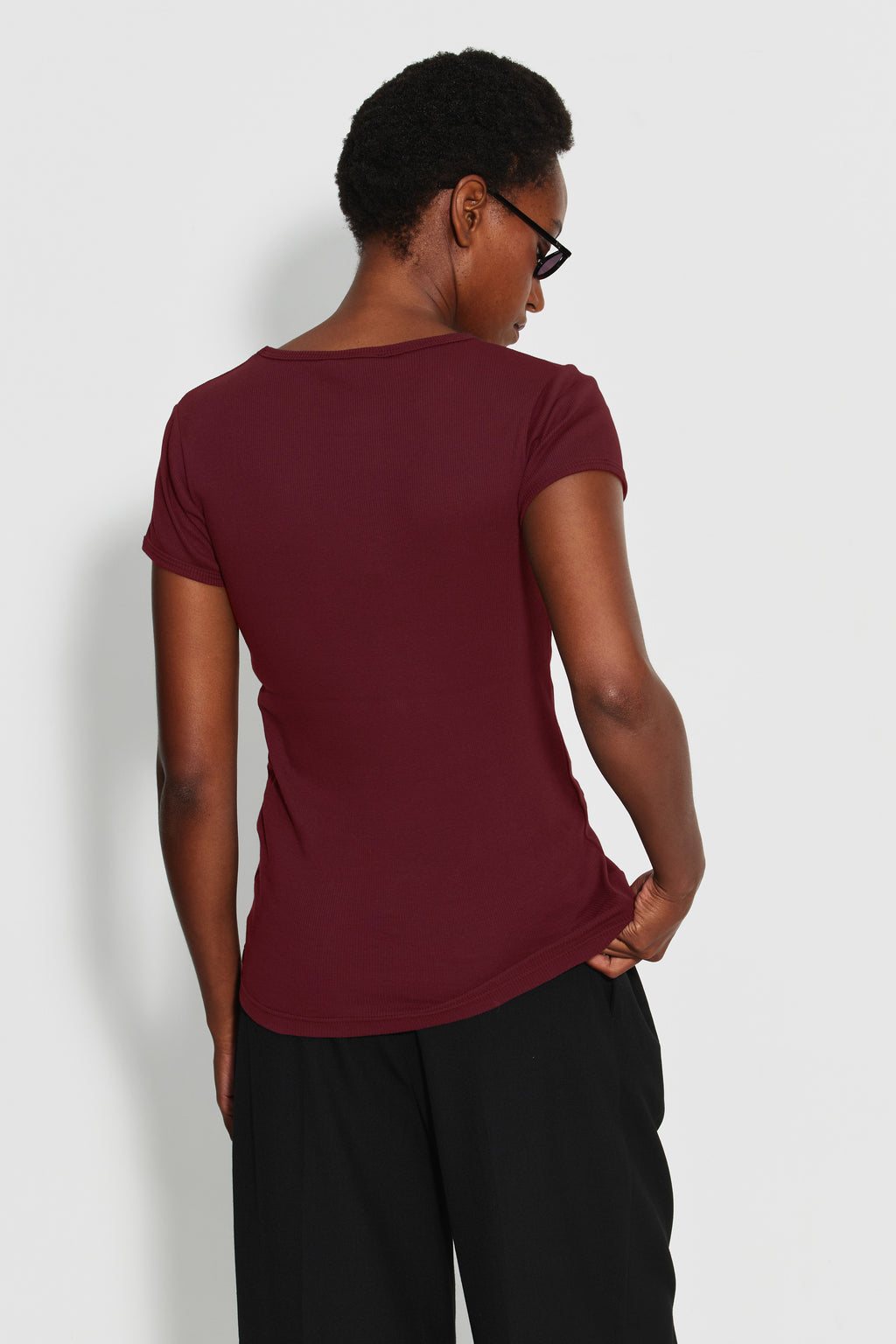 Maillot de corps femme bordeaux en coton pima côtelé à manches courtes, porté de dos avec un pantalon noir, la tête tournée sur le côté.