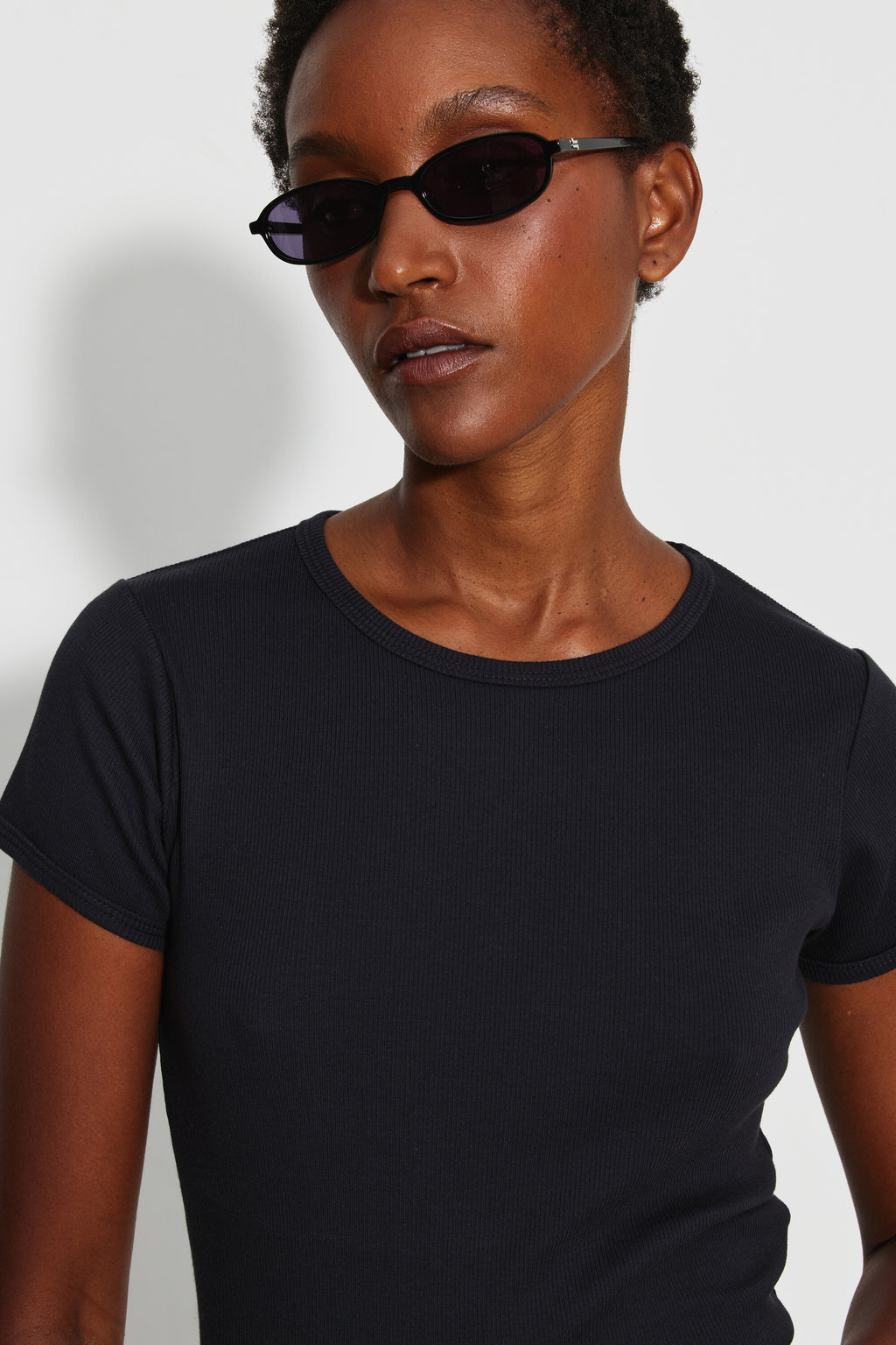 Maillot de corps femme noir à manches courtes en coton Pima côtelé, portrait rapproché montrant le col rond et les manches, avec des lunettes de soleil.