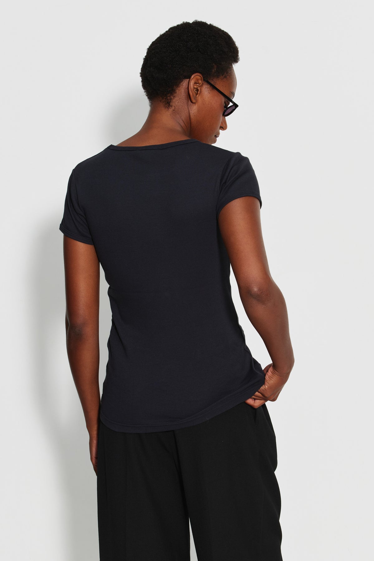Maillot de corps femme noir à manches courtes en coton Pima côtelé, porté de dos avec un pantalon noir, une main dans la poche.