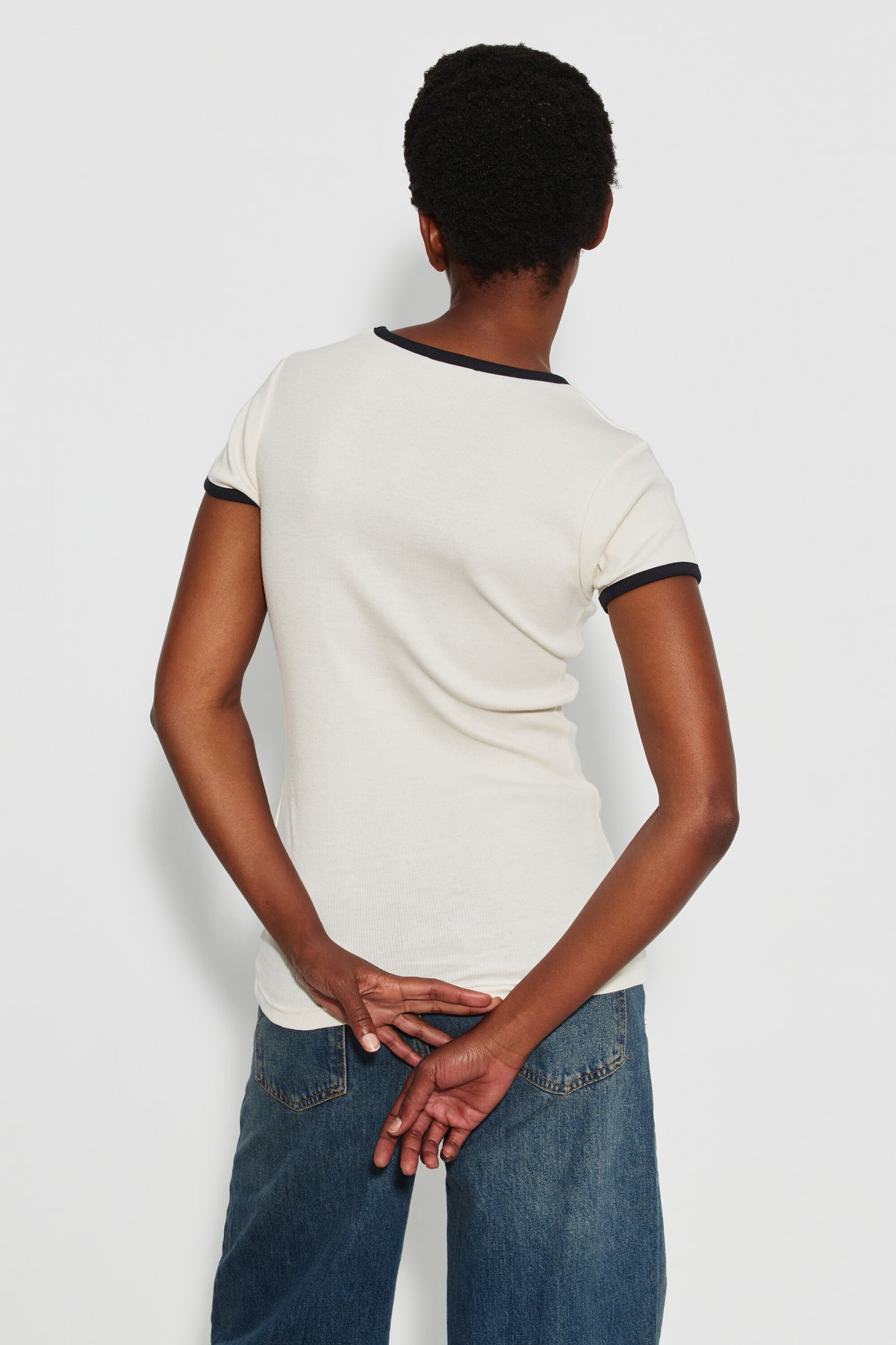 Maillot de corps femme bicolore écru et noir à manches courtes en coton Pima côtelé, porté de dos avec un jean bleu, les mains derrière le dos.