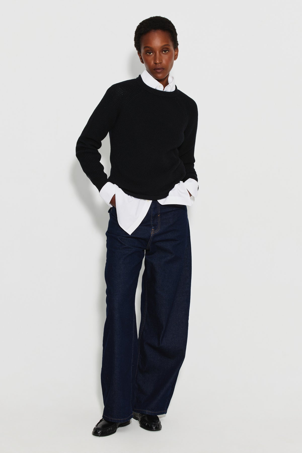 Pull femme noir en laine mérinos à manches raglan, porté sur une chemise blanche avec un pantalon bleu foncé, vue de face en pied. Made in France.