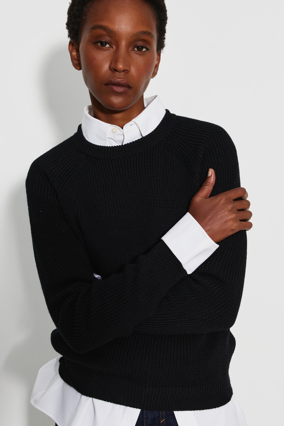 Pull femme noir en laine mérinos à manches raglan, portrait rapproché montrant le col rond et la maille, porté sur une chemise blanche, les bras croisés.