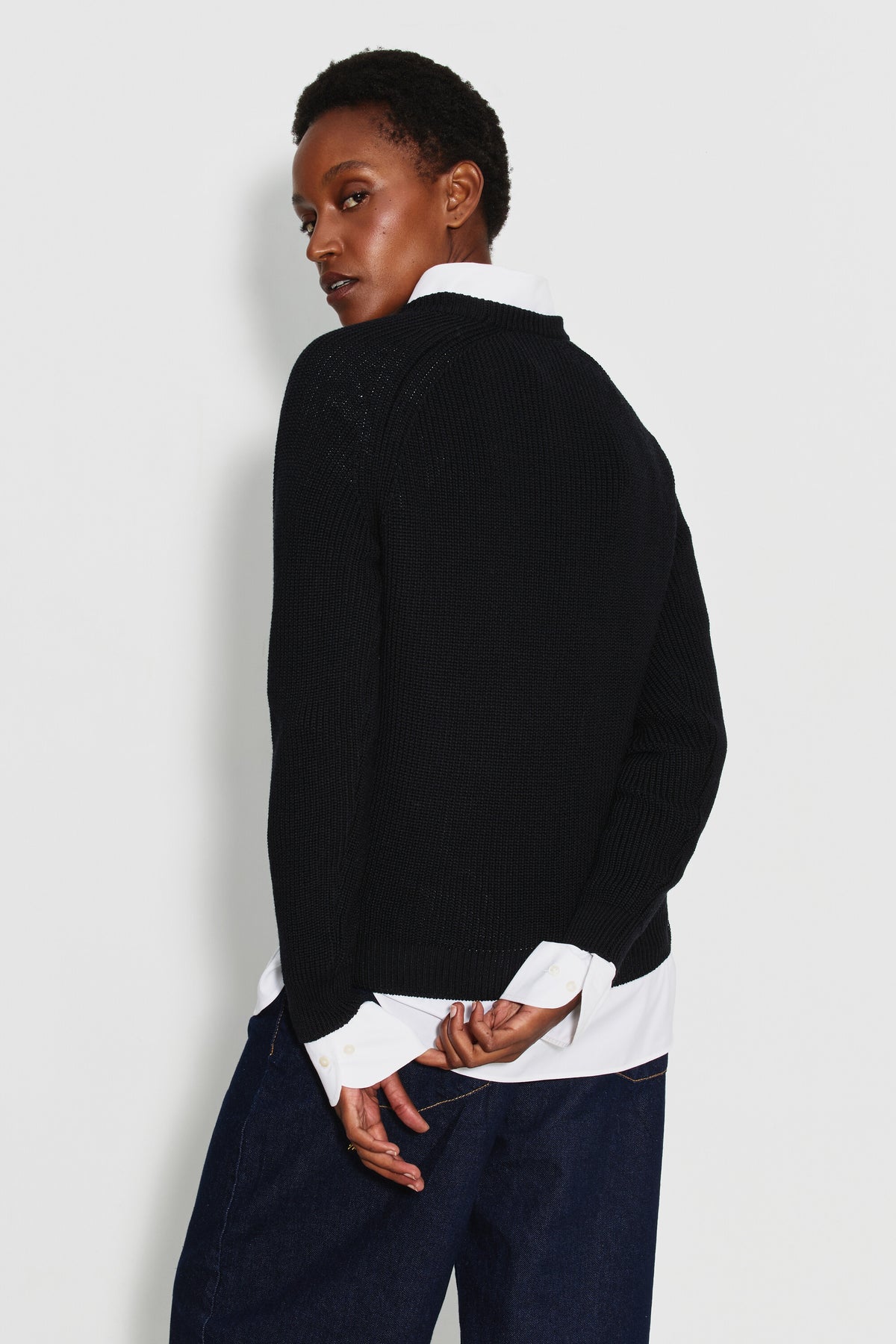 Pull femme noir en laine mérinos à manches raglan, porté de trois quarts dos sur une chemise blanche, la tête tournée vers l’objectif.