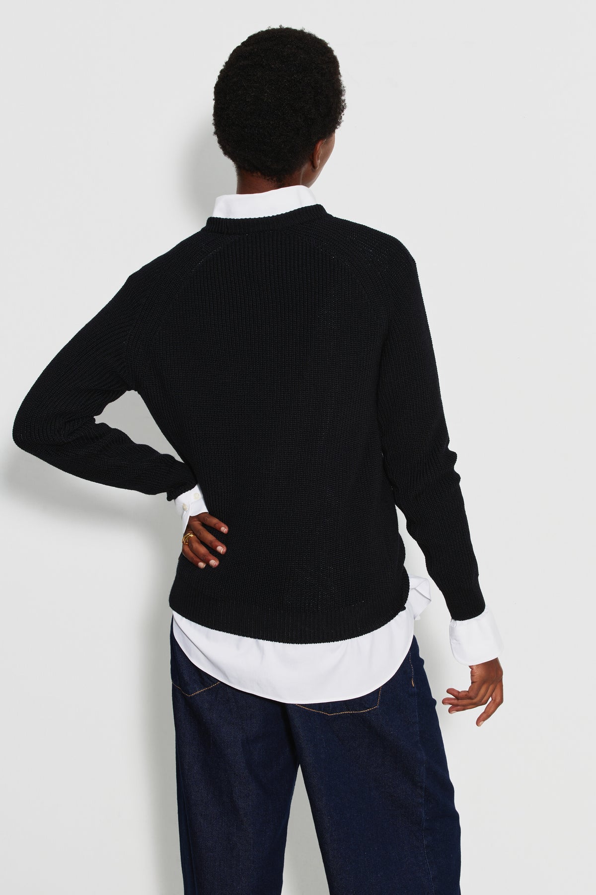Pull femme noir en laine mérinos à manches raglan, porté de dos sur une chemise blanche avec un pantalon bleu foncé, une main sur la hanche.