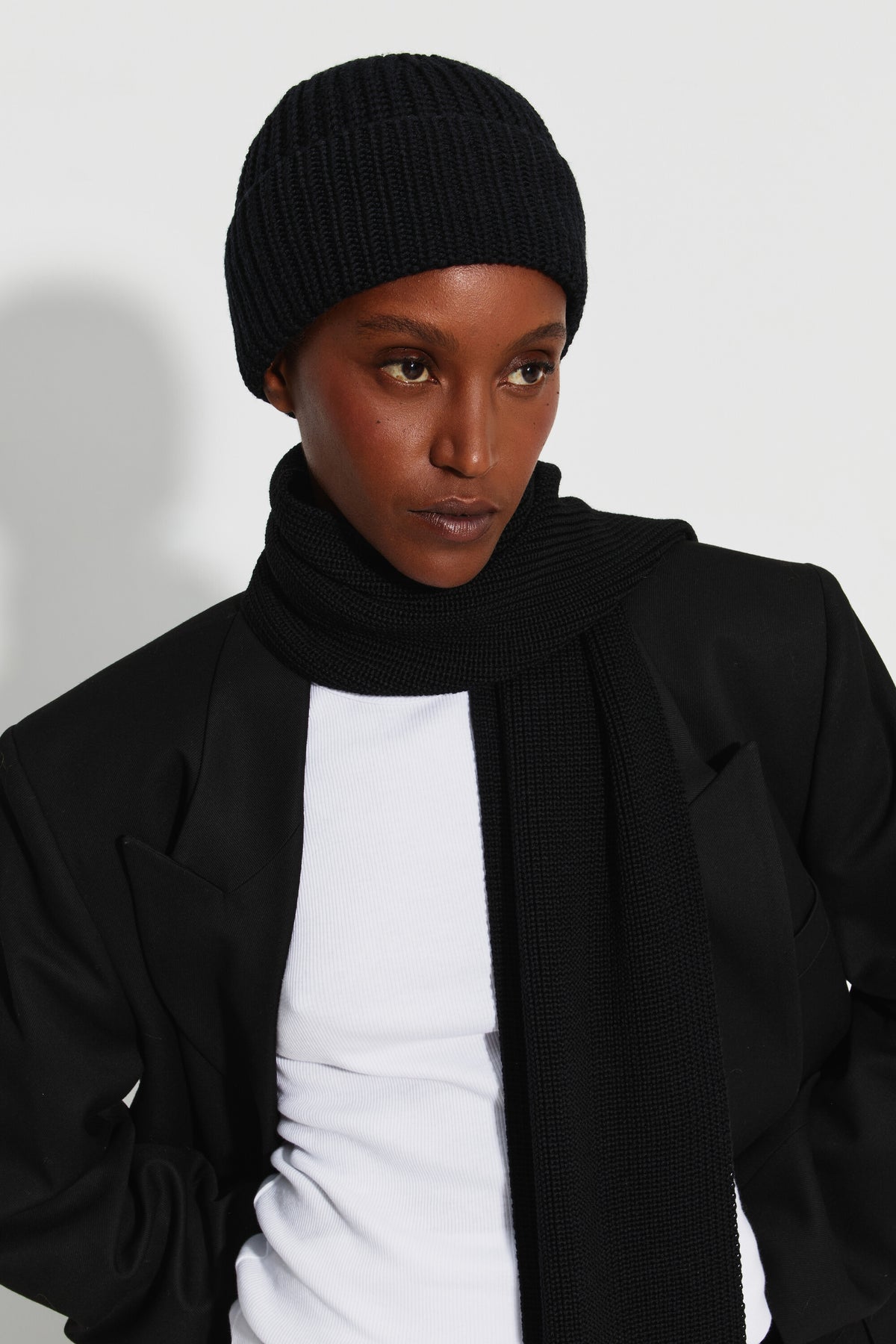 Bonnet femme noir en laine mérinos, porté de trois quarts avec une écharpe noire, un manteau noir et un haut blanc.