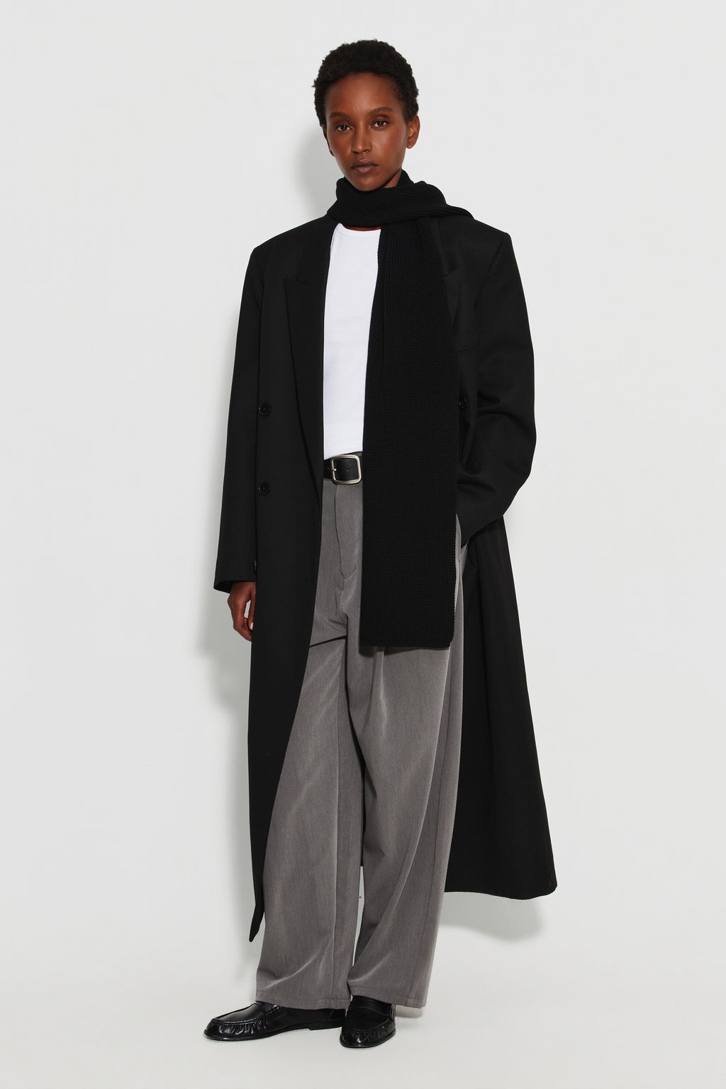 Écharpe femme noire en laine mérinos, portée avec un manteau noir, un haut blanc et un pantalon gris, vue de face en pied.