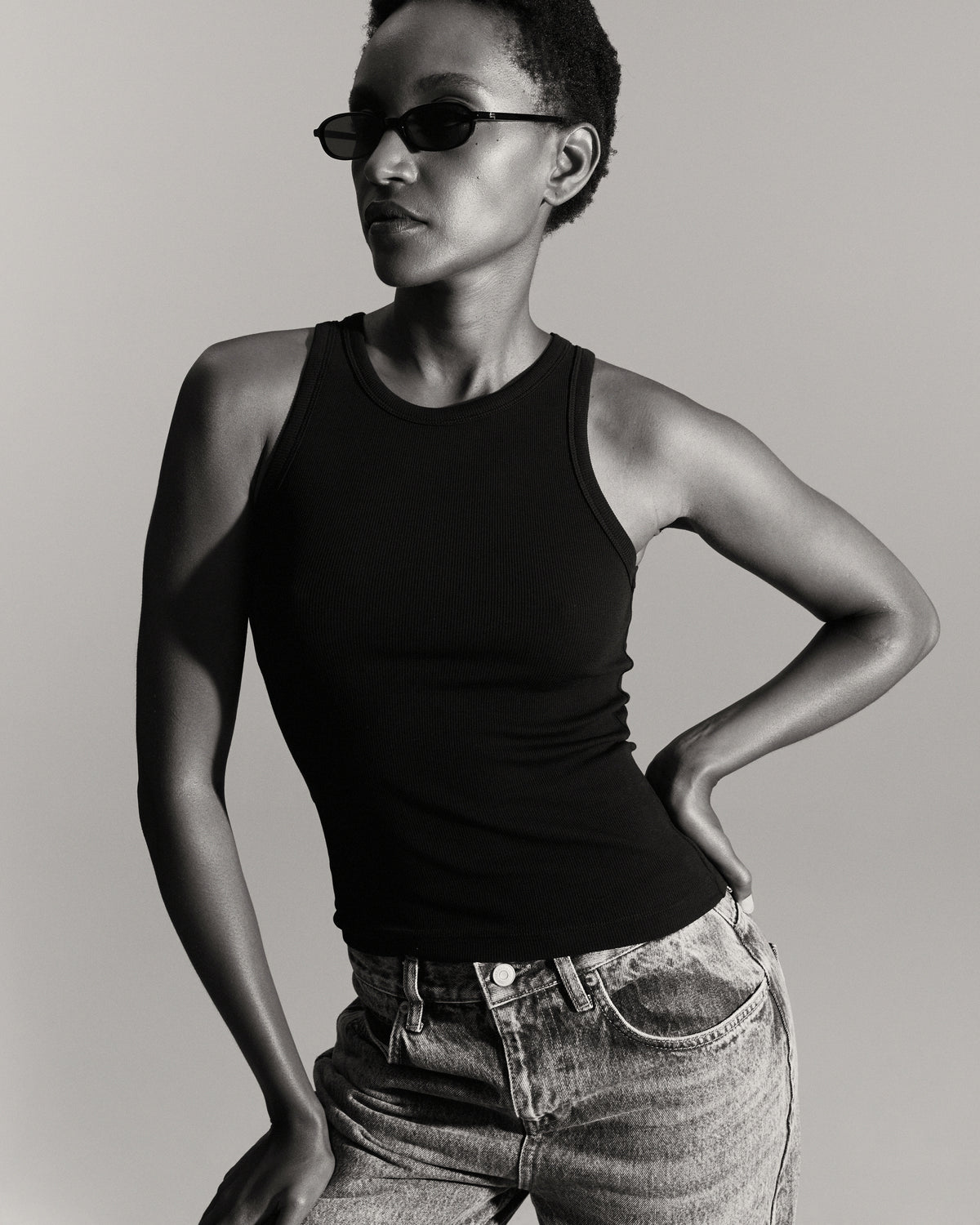 Débardeur Top Marcel noir femme, porté avec un jean et des lunettes de soleil, une main à la taille, photo en noir et blanc.