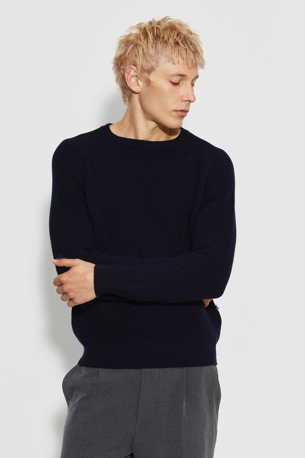 Pull homme bleu marine en laine mérinos à manches raglan, porté de face avec un pantalon gris, les bras croisés et la tête tournée sur le côté. Made in France.