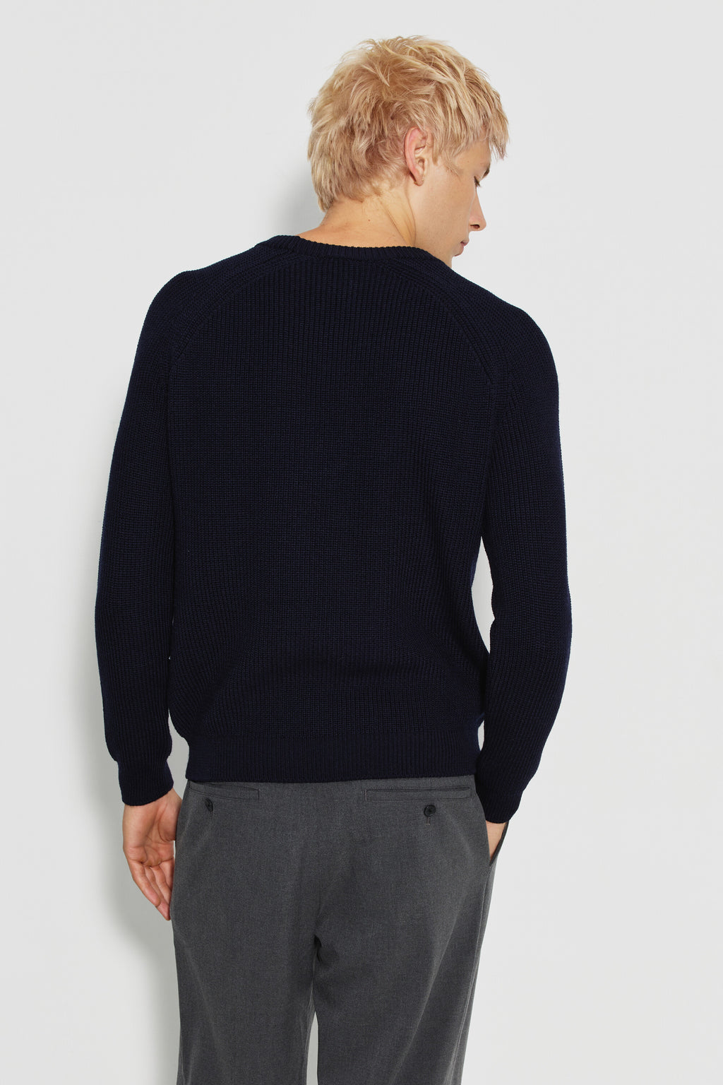Pull homme bleu marine en laine mérinos à manches raglan, porté de dos avec un pantalon gris, la tête tournée sur le côté.