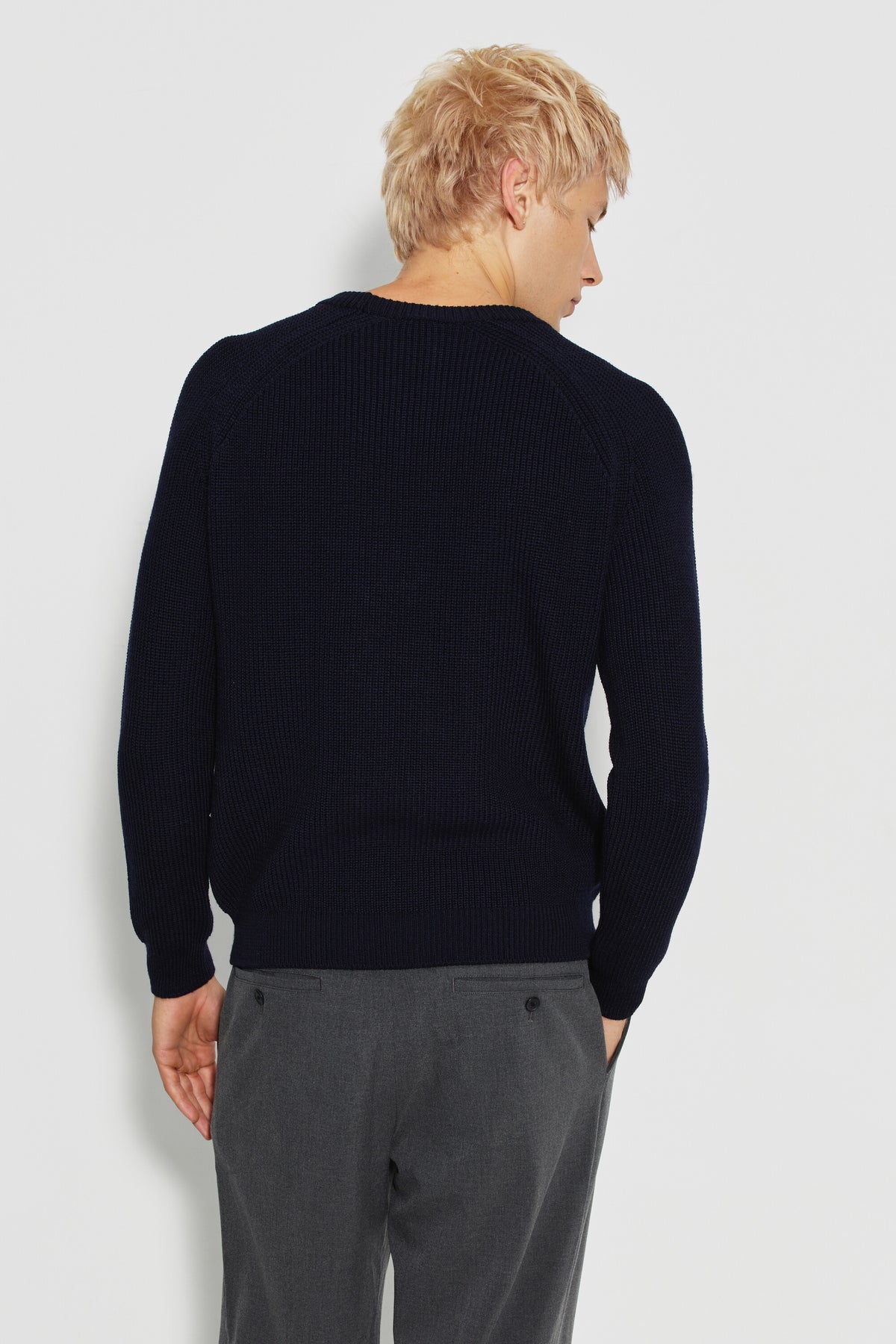 Pull homme bleu marine en laine mérinos à manches raglan, porté de dos avec un pantalon gris, la tête tournée sur le côté.