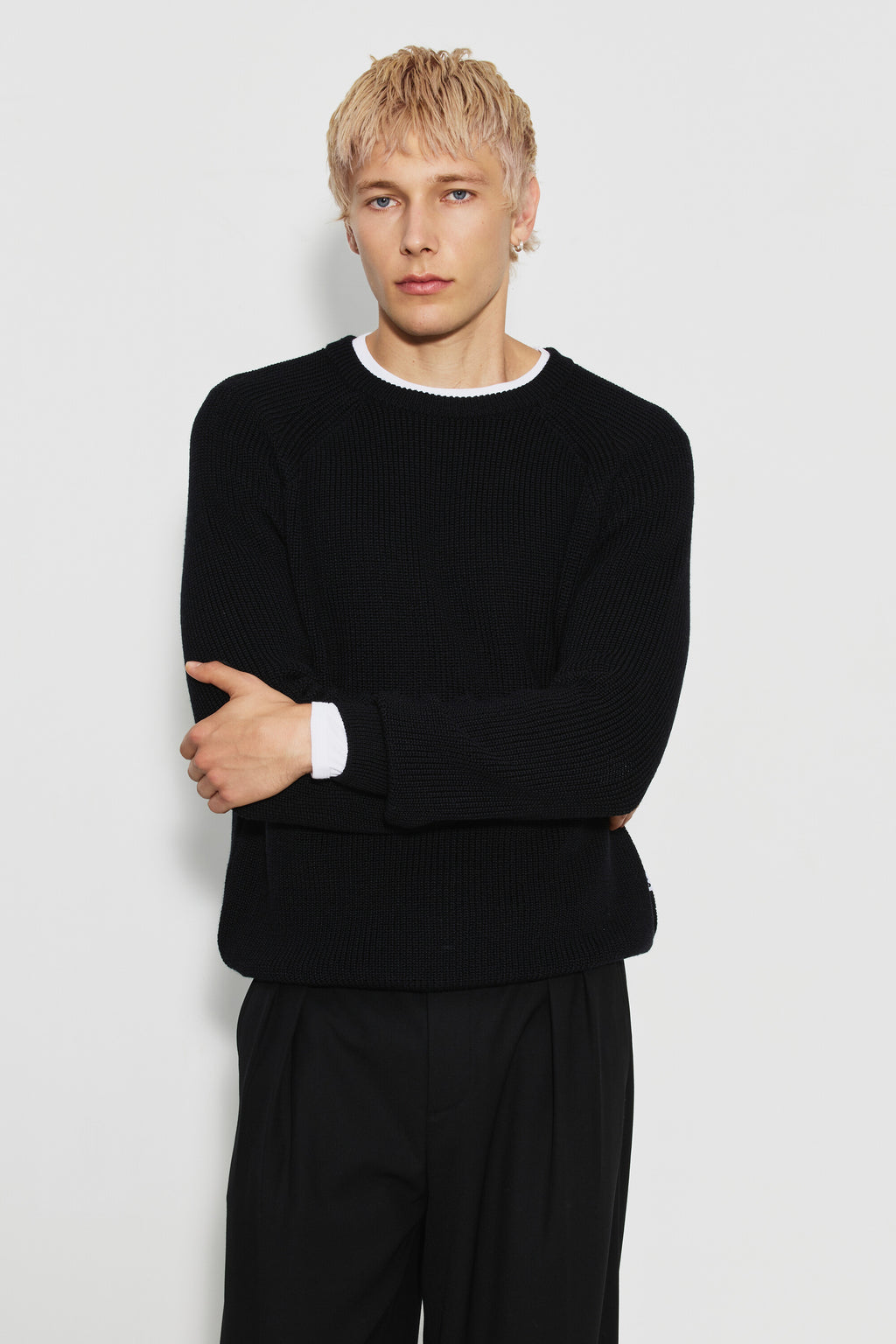 Pull homme noir en laine mérinos à manches raglan, porté de face avec un pantalon noir, les bras croisés et un sous-vêtement blanc visible au col. Made in France.