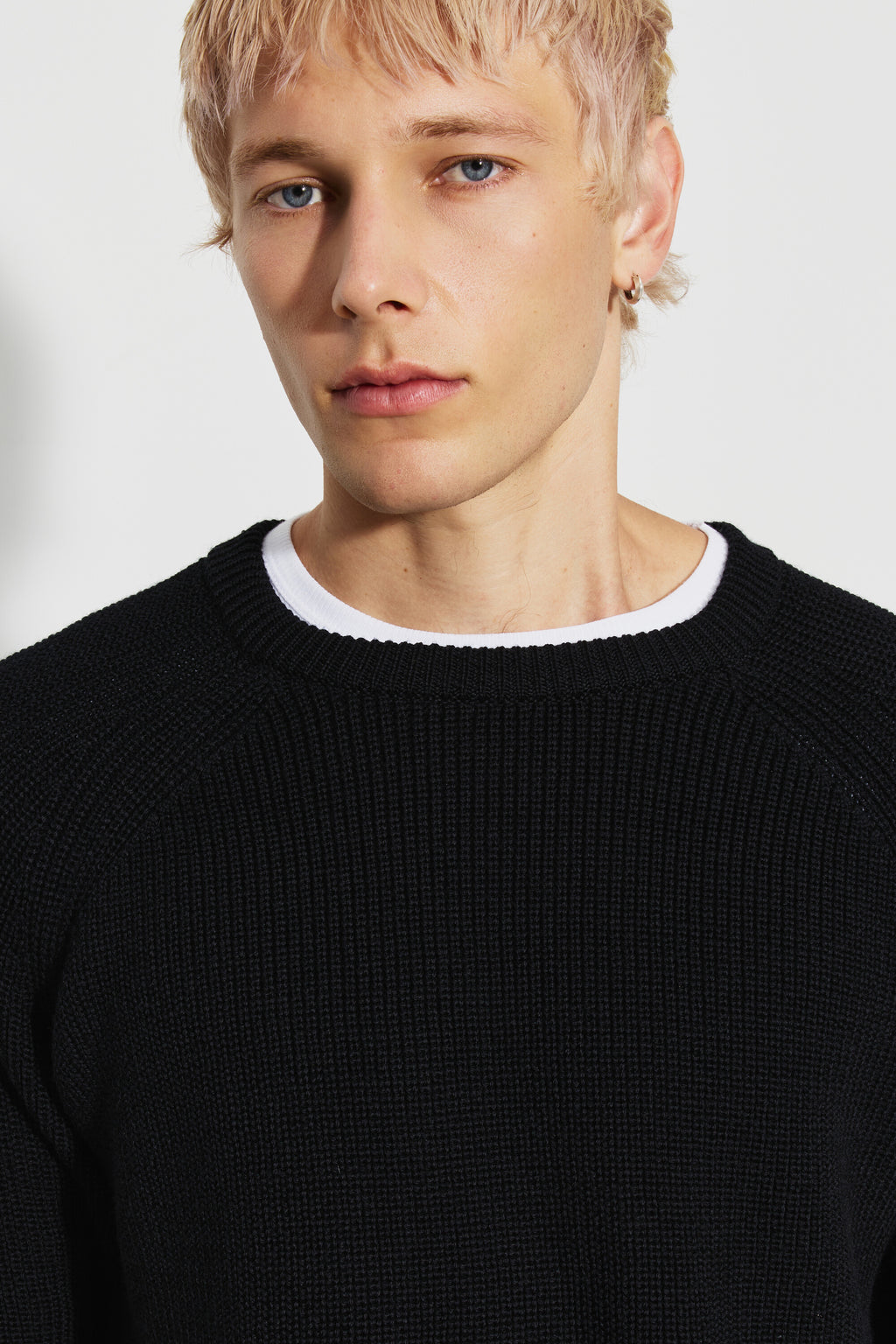 Pull homme noir en laine mérinos à manches raglan, portrait rapproché montrant le col rond et la maille perlée, avec un sous-vêtement blanc visible au col.