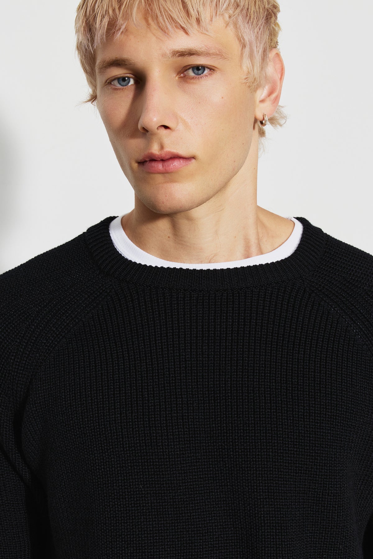 Pull homme noir en laine mérinos à manches raglan, portrait rapproché montrant le col rond et la maille perlée, avec un sous-vêtement blanc visible au col.