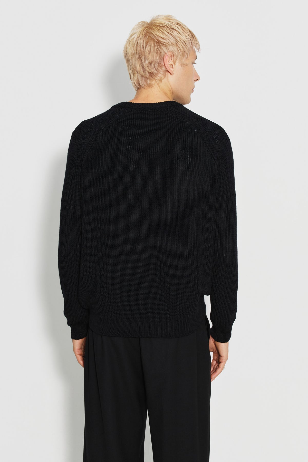 Pull homme noir en laine mérinos à manches raglan, porté de dos avec un pantalon noir, la tête tournée sur le côté.