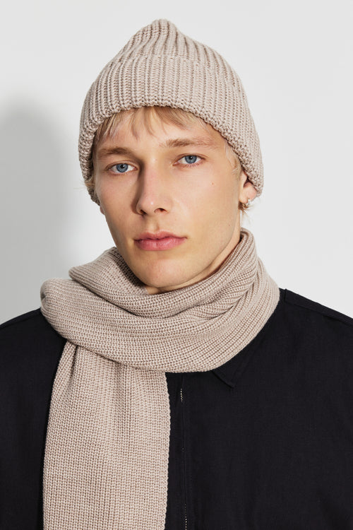 Bonnet homme beige en laine mérinos, portrait de face montrant le revers côtelé, avec une écharpe beige et un blouson noir. Made in France.