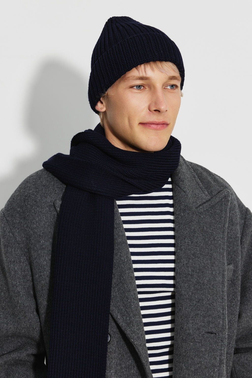 Bonnet homme bleu marine en laine mérinos, portrait de trois quarts avec une écharpe assortie, un manteau gris et une marinière. Made in France.
