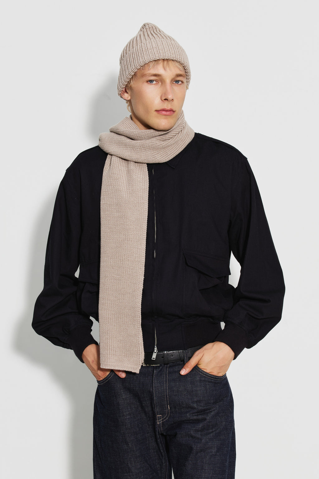 Écharpe homme beige en laine mérinos, portée autour du cou avec un pan sur la poitrine, un bonnet assorti et un blouson noir. Made in France.