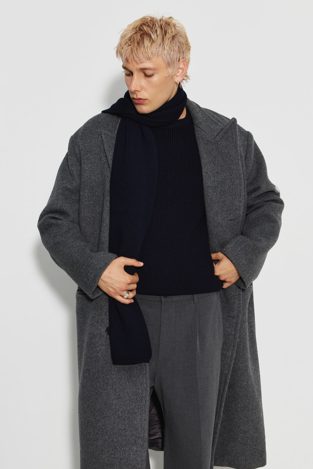 Écharpe homme bleu marine en laine mérinos, portée avec un manteau gris, les mains tenant les revers du manteau et la tête baissée.