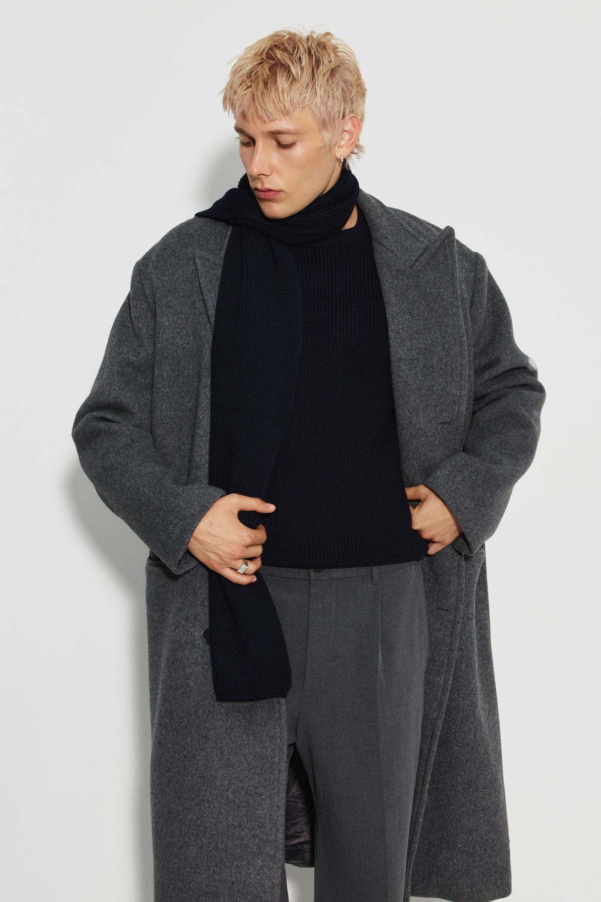 Écharpe homme bleu marine en laine mérinos, portée avec un manteau gris, les mains tenant les revers du manteau et la tête baissée.
