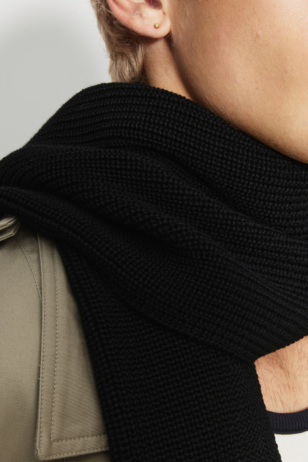 Écharpe homme noire en laine mérinos, gros plan de la maille de l’écharpe enroulée autour du cou, avec un trench beige.