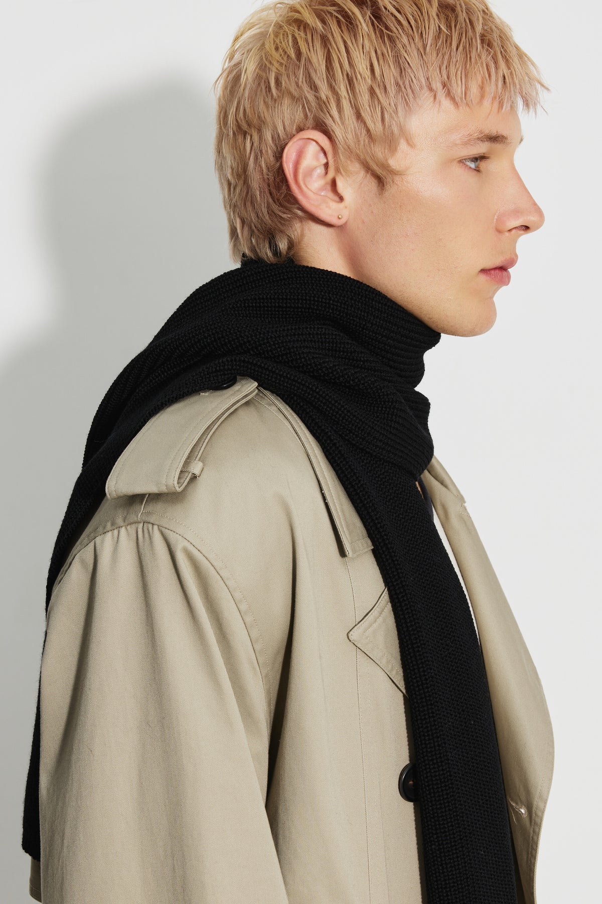 Écharpe homme noire en laine mérinos, portée enroulée autour du cou avec un trench beige, vue de profil.