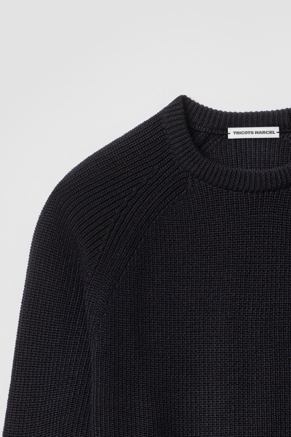Pull homme et femme noir en laine mérinos à manches raglan, détail du col rond, de l’épaule raglan et de la maille perlée. Made in France.